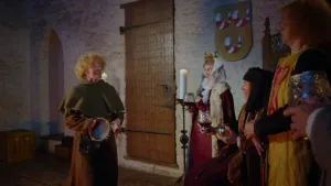 Een foto van een koning op een stoel rechts in beeld en een koningin en een andere man die ernaast staan. Links in beeld is een man te zien in een bruin pak met groene doek om zijn nek. De man heeft een tamboerijn, een trommel met belletjes eraan, vast. 