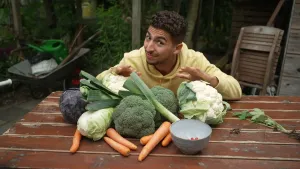 Een foto van presentator Nizar die buiten aan een tafel zit waar allemaal groentes op liggen, waaronder wortels, bloemkool, broccoli en prei.