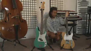 Een foto van presentator Eva die op de grond zit tussen twee basgitaren in. Links staat er een cello en op de achtergrond staan er elektrische piano's.