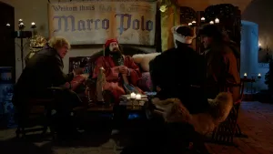 Een foto van een man verkleed als Marco Polo in een rood pak en met een rode muts. Achter hem hangt een krantenartikel waarop staat: "Welkom thuis Marco Polo". Ernaast zit een man met een soort microfoon. Ervoor is de rug van twee andere mensen te zien.