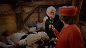 Een foto van een man verkleed in een rood pak met een rode hoed die een soort microfoon in zijn handen heeft. Hij stelt een vraag aan een man in een zwart pak. Erachter en ernaast liggen zieke mensen op een bed van stro. Ze hebben de ziekte de pest.