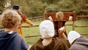 Een foto van een man die met zijn handen en hoofd voor straf door een houten schandpaal gestoken is. Er staat een verslaggever met een soort microfoon naast. De man heeft witte stukken ei in zijn haar en Iemand vooraan wil een wit, rot ei naar hem gooien.