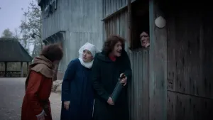 Een foto met het hoofd en de handen van een als dokter Balavignus verklede man, rechts zichtbaar achter het luik van zijn woning. Naast de woning staat een man met een opgerold document in handen. Eén man en één vrouw kijken naar achteren en naar links.