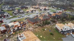 Een foto van een straat met door een tornado kapotgemaakte huizen, waarvan de daken deels of helemaal afgewaaid zijn. Hout en spullen liggen op de groene grasvelden ervoor. Op de weg erachter zijn politieauto's en een stukje van een brandweerauto te zien.