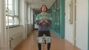 Een foto van presentator Pascal die in een gang staat met drum stokjes in zijn handen die hij gekruist vasthoudt. Voor hem staat een kleine drum.