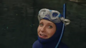 Een foto van presentator Janouk met een duikpak aan en een duikbril en snorkel op. Achter haar is water te zien.