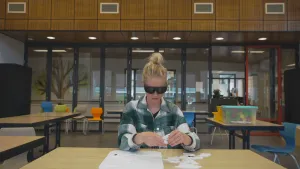 Een foto van presentator Janouk die aan tafel zit en met een spel bezig is terwijl ze een zonnebril op heeft waar je niet doorheen kan kijken.