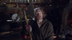 Een foto van een man die in een donkere, houten schuur staat waar bruine kettingen aan de muur hangen. De man kijkt met opengesperde ogen naar rechts. Hij heeft in handen een houten handvat met een metalen hakbijl eraan. Hij draagt een bruin kostuum.