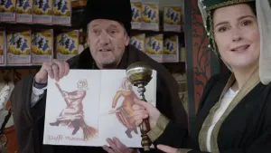 Een foto van een man die een boek vastheeft met twee plaatjes erin van heksen op bezemstelen. De man heeft een bult op zijn wang en draagt een bruin pak en zwarte hoed. Ernaast staat een vrouw in zwart met bruin kostuum en een soort microfoon in handen.