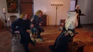 Een foto met daarop links twee mannen die op een stoel zitten met gekruisde benen. Ze kijken naar rechts waar een vrouw verkleed als Jacoba van Beieren zit in een donkerkleurig kostuum met witte mouwen en met een witte, doorzichtige sluier op haar hoofd. 