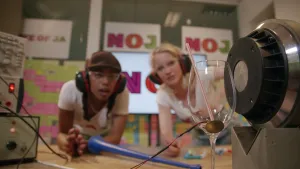 Een foto van Eva en Janouk achter een tafel met gehoorbeschermers en een veiligheidsbril. Links staan geluidsapparaten met druk- en draaiknopjes, rechts een luidspreker naast een wijnglas met plastic rietje erin en vastgeplakte rode en zwarte draden erop.