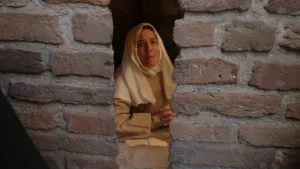 Een foto van een vrouw verkleed als Non Suster Bertken in lichtbruine kleding en witte doek om haar hoofd voor een tafel met een boek. Ze zit achter een spleet van een muur met roodbruine stenen. Ze zit met gevouwen handen te bidden en kijkt serieus. 