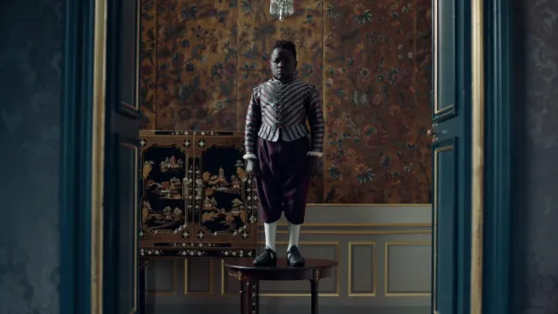 Een foto van een jongen van kleur, die op een tafeltje staat in een kamer. Zijn kleren en de kamer zien eruit als uit de zeventiende eeuw. De kamer is versierd met veel goudkleurige verf en behang. 