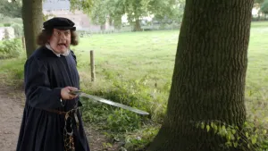 Een foto van een man verkleed als François Villon die met opengesperde ogen en open mond naar rechts kijkt. Hij heeft een zilverkleurig zwaard vast en wijst ermee naar rechts. De man draagt een donker pak en een pet. Hij staat op een pad naast een boom.