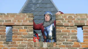 Een foto van een man verkleed als Jan van Schaffelaar in een harnas en met helm op. Hij staat achter een stenen muur voor een zwarte toren en kijkt naar beneden. In de muur zit een rechthoekige opening, waar de man de beide kanten van vasthoudt. 