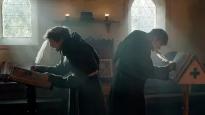 Een foto van twee mannen in een pei een bruin pak met capuchon. Ze staan met de rug naar elkaar met een veer in een boek te schrijven. De kamer is verlicht door twee ramen met zwarte driehoeken ervoor. Ze zijn monniken, die samen wonen en geloven in god.