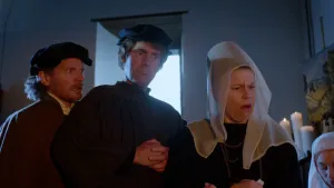 Een foto van drie mensen in een donkere kamer met klein raam. De mensen kijken verbaast met grote ogen, twee met hun mond open één met de mond stijf dicht. Twee mannen dragen een donkerkleurige pet, één vrouw een hoge hoed met een witte sluier erover. 