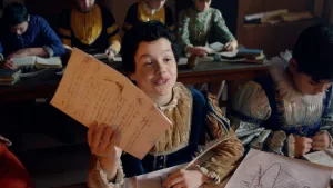 Een foto van een jongen in een schoolklas achter een bureau in een blauw met bruin pak. In één hand heeft hij een papier met tekst, in de andere een witte veer, waar mensen vroeger mee schreven. Naast zijn arm ligt een deel van een tekening op papier.