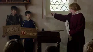Een foto van een man in een rood, middeleeuws pak voor een klas die wijst naar twee jongens. De man heeft een wit bord met een ketting vast. De jongens staan tegen de muur met houten borden om hun nek, één van een getekende ezel en één met "Domoor" erop.