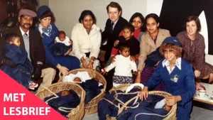 Een foto van ongeveer tien volwassen mensen en enkele kinderen. De mensen hebben verschillende huidskleuren. Op de grond staan vier wiegjes met donker gekleurde baby's erin. Linksonder in beeld staat de tekst 'Met lesbrief'.