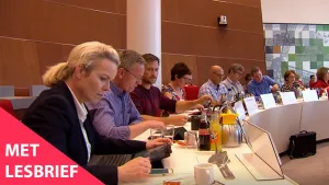Een foto van een groep mensen die aan een lange tafel zitten. De tafel loopt in een bocht en de mensen zitten allemaal aan dezelfde kant. Sommigen hebben een laptop bij zich en er staat drinken op tafel. Linksonder in beeld staat de tekst 'Met lesbrief'.