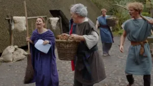 Een foto van vier mensen in Middeleeuwse kleding. Een vrouw draagt een bezem en een man draagt een mand. Ze lopen op straat. Op de achtergrond zie je een woning met een rieten dak dat doorloopt tot aan de grond. 