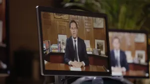 Een foto een beeldscherm met Mark Rutte erop die een toespraak houdt terwijl hij recht voor de camera aan een tafel zit. Op de achtergrond is er een nog een beeldscherm te zien met hetzelfde beeld.