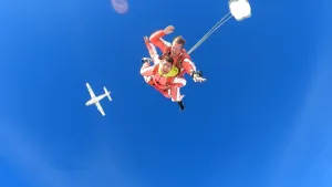 Een foto van presentator Nizar die een parachute sprong maakt waar hij vastzit aan een ervaren parachute springer. Op de achtergrond is de onderkant van het vliegtuig te zien waar ze uit zijn gesprongen.