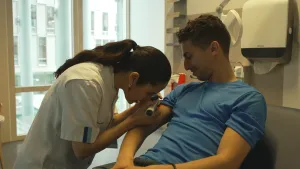 Een foto van presentator Nizar die bij een huiddokter in de stoel zit. De huiddokter kijkt met speciaal gereedschap naar de huid op de bovenarm van Nizar.