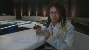 Een foto van presentator Eva die voor de camera zit in een zaal van een ambassade. Voor haar ligt er een stapel papieren op tafel.