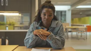 Een foto van presentator Sosha die aan een tafel zit in een lokaal van een school. Ze zit recht voor de camera en op de achtergrond is er een grote gang te zien.