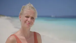 Een foto van presentator Anouk die in de camera kijkt terwijl ze op een strand staat van een tropisch gebied. 