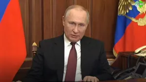 Een foto van de Russische leider Poetin. Hij heeft een net pak aan en zit aan een bureau met naast hem twee vlaggen en twee telefoons. Hij is iets aan het vertellen. 