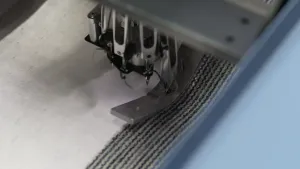 Een foto van een stuk wit stof, dat onder een machine ligt. Aan de zilverkleurige arm van de machine hangen kleine haakjes of naalden. De machine is bezig de zwartgrijze tapijtstof op de witte stof te weven.