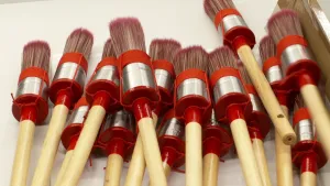 Een foto met zestien kwasten erop. Het zijn verfkwasten om muren of andere dingen mee te verven. De kwasten hebben een lichtbruin houten handvat dat aan een zilverkleurig deel met rode stof eromheen zit. De lange, rechtopstaande haartjes zitten bovenop.