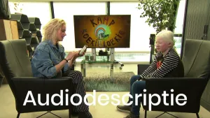 Een foto van Janouk en Koen die ieder op een stoel zitten en ervoor het woord "Audiodescriptie", horen wat er in bewegende beelden gebeurt. Janouk heeft haar mobiele telefoon vast en kijkt ernaar. Op de tv erachter staat de tekst "Kamp Koekieloekie".