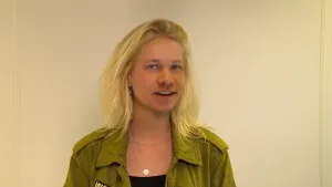 Een foto waarop een beeldbewerking te zien is. In het beeld staat Janouk voor een witte muur. Te zien zijn Janouk's lichtblonde haar, groene vest, zwart t-shirt en kettingen om haar nek. Het gezicht is alleen niet van Janouk, maar van een lachende Pascal.