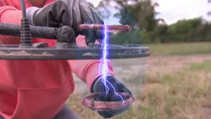 Een foto van het zwarte, ronde blad van een metaaldetector en de stekker die er bovenop uitsteekt. Iemand houdt twee spoelen rood metaalraad boven en onder het blad. Daartussen is een paars met wit magnetisch veld te zien, dat lijkt op een bliksemflits.