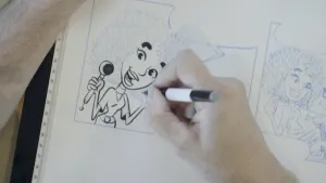 Een foto van een wit vel papier met daarop tekeningen van een stripverhaal in lichtblauwe lijnen en twee nog lege tekstballonnen. Een hand is de linkertekening met een zwarte stift over aan het trekken. De tekening is van een vrouw met een microfoon. 