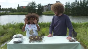 Een foto van Eva en Florian op een groen grasveld aan de waterkant. Op de tafel ervoor ligt een stapel takken en twee plastic zakken met dingen erin. Rechts staat een opgezette vogel, een meerkoet, zwart met witte snavel. Eva houdt beide handen omhoog.  