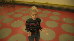 Een foto van Janouk die in een ondergronds gebouw staat en haar handen met de handpalmen naar voren houdt. Ze kijkt omhoog naar de camera en staat op een rode vloer, met grijze cirkels erop. Onder de cirkels ligt kernafval van een kerncentrale opgeslagen.