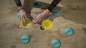 Een foto van een houten vloer met zes groenblauwe rondjes met het woord 'Uranium", zes rode rondjes met het woord "Afval" en zes zwarte rondjes met de letter "N", voor Neutron. Om twee groepjes rode en zwarte rondjes staat een doorzichtig, geel rondje.