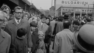 Een foto in zwartwit van tientallen mensen op straat in een stad. Een deel van de mensen staat in een rij. Op de achtergrond zie je huizen, winkels en fabrieken.
