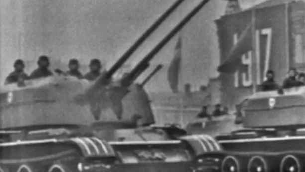 Een zwart-witfoto van twee oorlogstanks die voorbij een groot gebouw rijden. Bovenin de tanks zijn mensen te zien. Op het gebouw staat in grote cijfers 1917.