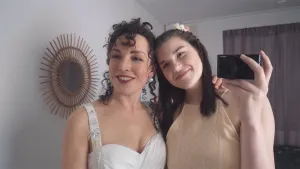 Een foto van Lana in een bruine jurk met witte bloemen erop rechts en haar Moeder in een witte trouwjurk links, die allebei lachen. Ze kijken naar links en Lana neemt een foto in een niet-zichtbare spiegel met de zwarte fotocamera in haar linkerhand.