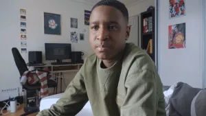 Een foto van Gino in zijn kamer. Gino kijkt verdrietig voor zich uit. Hij draagt een groene trui en achter hem zijn een stukje van zijn bed en zijn computerbureau met beeldscherm te zien. Hij vertelt zijn verhaal over seksueel misbruik via internet.