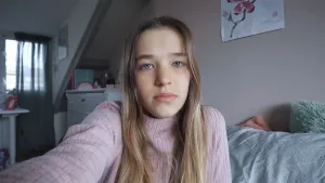 Een foto van Floor in haar kamer. Ze draagt een roze trui en heeft donkerblond haar dat langs haar schouders loopt. Floor kijkt verdrietig in de camera die ze zelf vastheeft voor haar gezicht. Ze vertelt haar verhaal over seksueel misbruik.