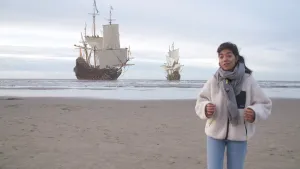 Een foto van een presentator op het strand. In het water zijn twee op de foto toegevoegde Nederlandse handelsschepen te zien. De schepen zijn van hout en hebben meerdere witte zeilen en Nederlandse vlaggen in de masten. 