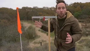 Een foto van een presentator in een bosgebied. Naast hem staat een Romeinse Groma, een soort liniaal om wegen recht te laten lopen. Het is een paal met vier grijze stroken in een plusvorm. Aan iedere strook hangt een klein, zwart gewicht aan een touwtje.