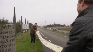 Een foto van twee mannen op een grasveld langs een autoweg. Ze zijn bezig een lange Romeinse wegenkaart uit te rollen. Aan de linkerkant staat een grijze mijlpaal, een ronde paal met de afstand naar en naam van de toekomstige plaats erop.